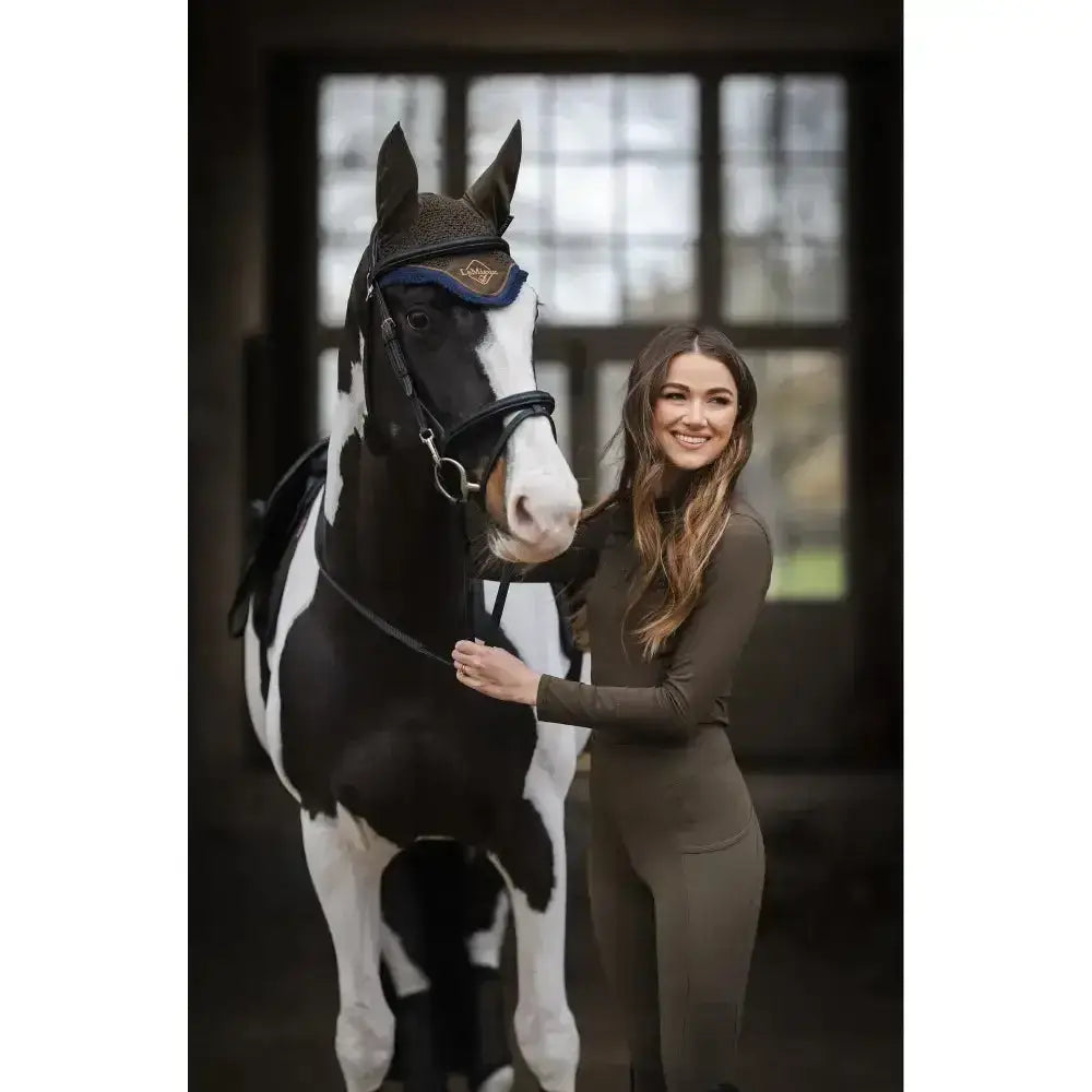 A black and white pinto horse wearing a lemieux classic fly hood alpine with a dark brown bridle with blue accents