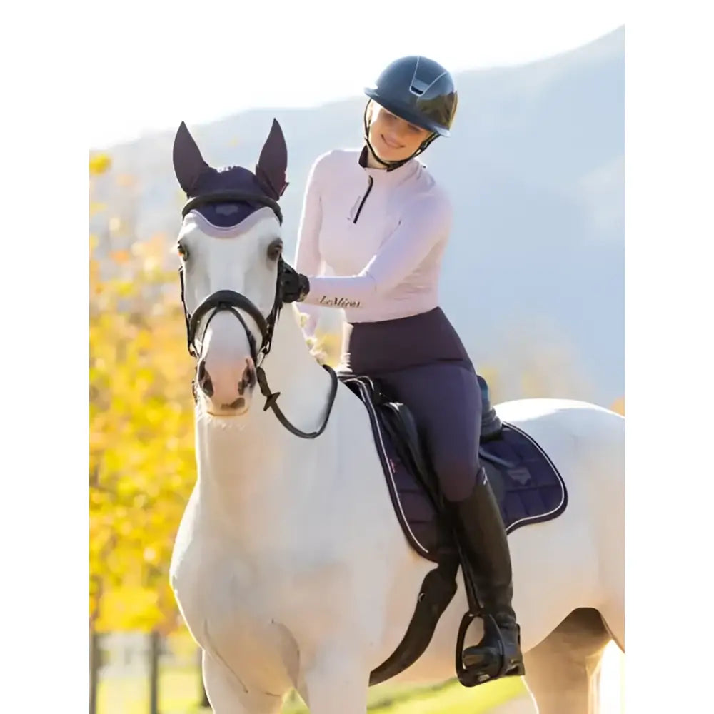 A white horse wearing a dark blue saddle pad with lemieux classique base layer lilac and black bridle and ear covers
