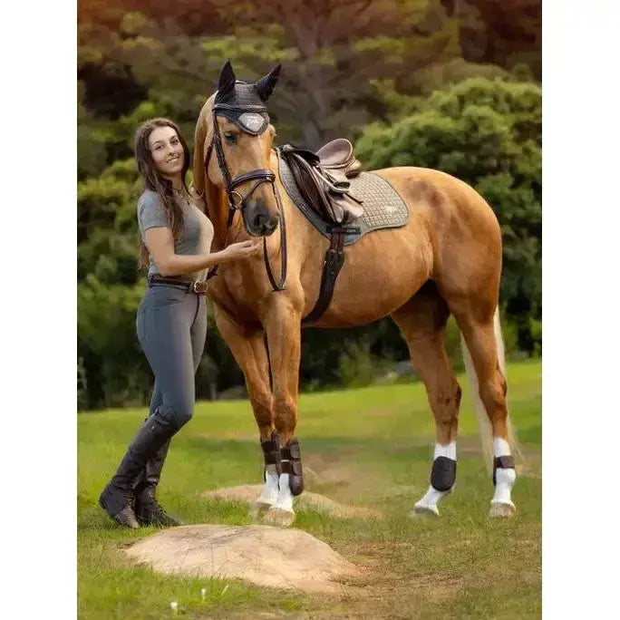 A tan horse wearing black protective gear and a lemieux earth close contact square moss saddle pad outdoors
