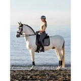 A white horse wearing a dark exclusive earth dressage saddle with black leg wraps and lemieux earth dressage square moss