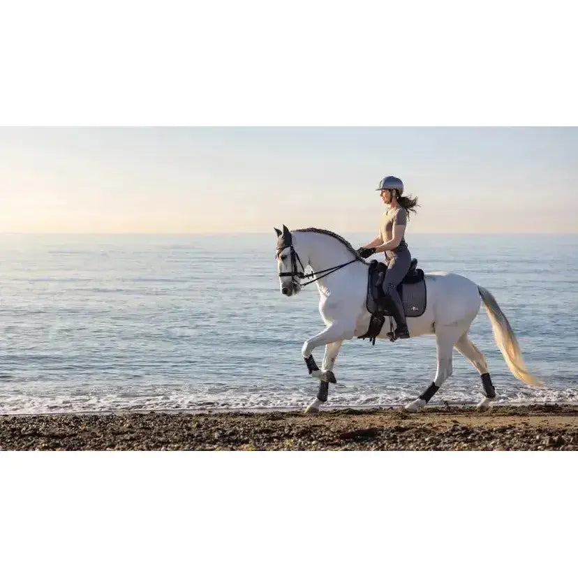 A white horse with black leg wraps and exclusive lemieux earth dressage saddle riding by shore in earth dressage square moss