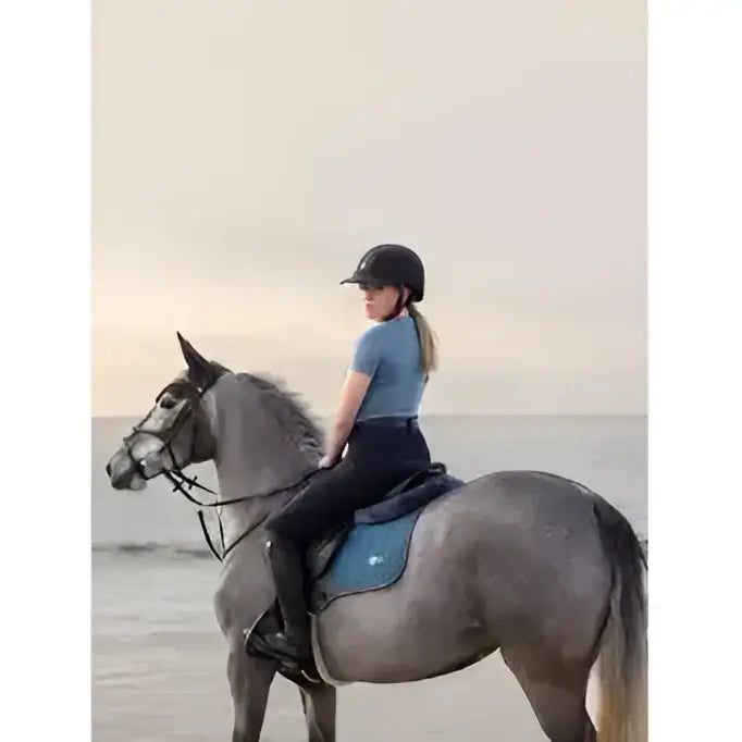 A young woman rides a gray horse wearing a lemieux earth gp square ocean saddle pad in blue and black riding gear