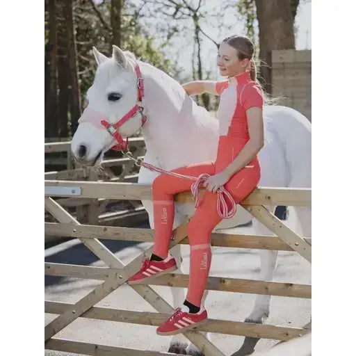 A woman in coral outfit wearing lemieux harlow competition socks dusty cora seated next to a white horse on a wooden fence