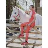 A woman in coral outfit wearing lemieux harlow competition socks dusty cora seated next to a white horse on a wooden fence