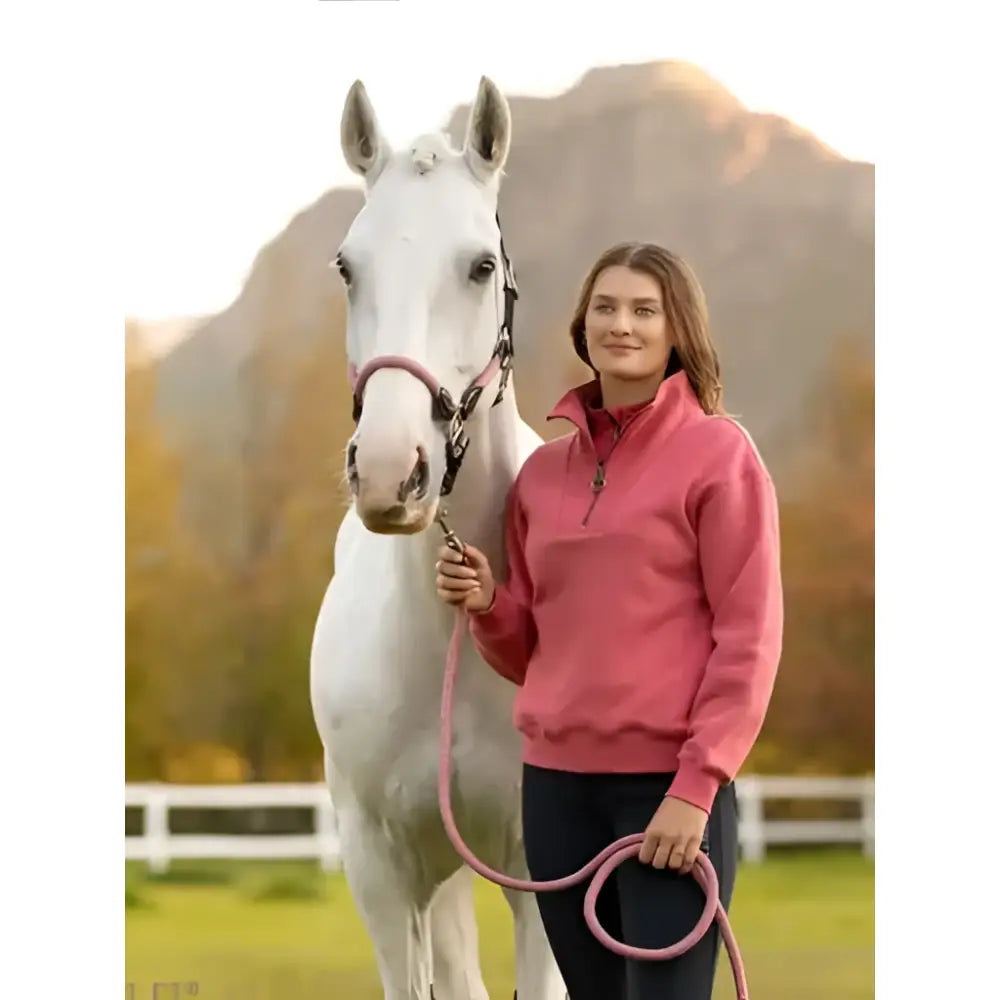 A woman stands beside a white horse wearing a lemieux keira quarter zip sweat cranberry pink fleece pullover holding the lead rope