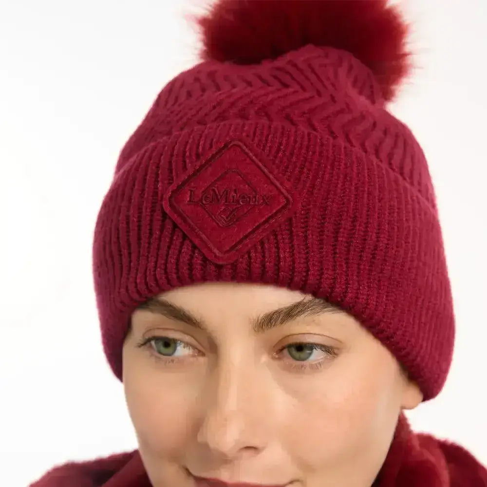 A maroon knitted lemieux lila beanie ember with a fluffy pom-pom and diamond-shaped embroidered patch