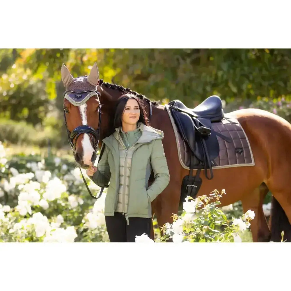 A woman in a light green quilted jacket stands beside a brown horse with a lemieux loire classic dressage square walnut saddle pad