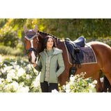A woman in a light green quilted jacket stands beside a brown horse with a lemieux loire classic dressage square walnut saddle pad