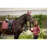 A horse wearing a lemieux loire fly hood orchid with matching maroon ear covers and a quilted pad under a black saddle