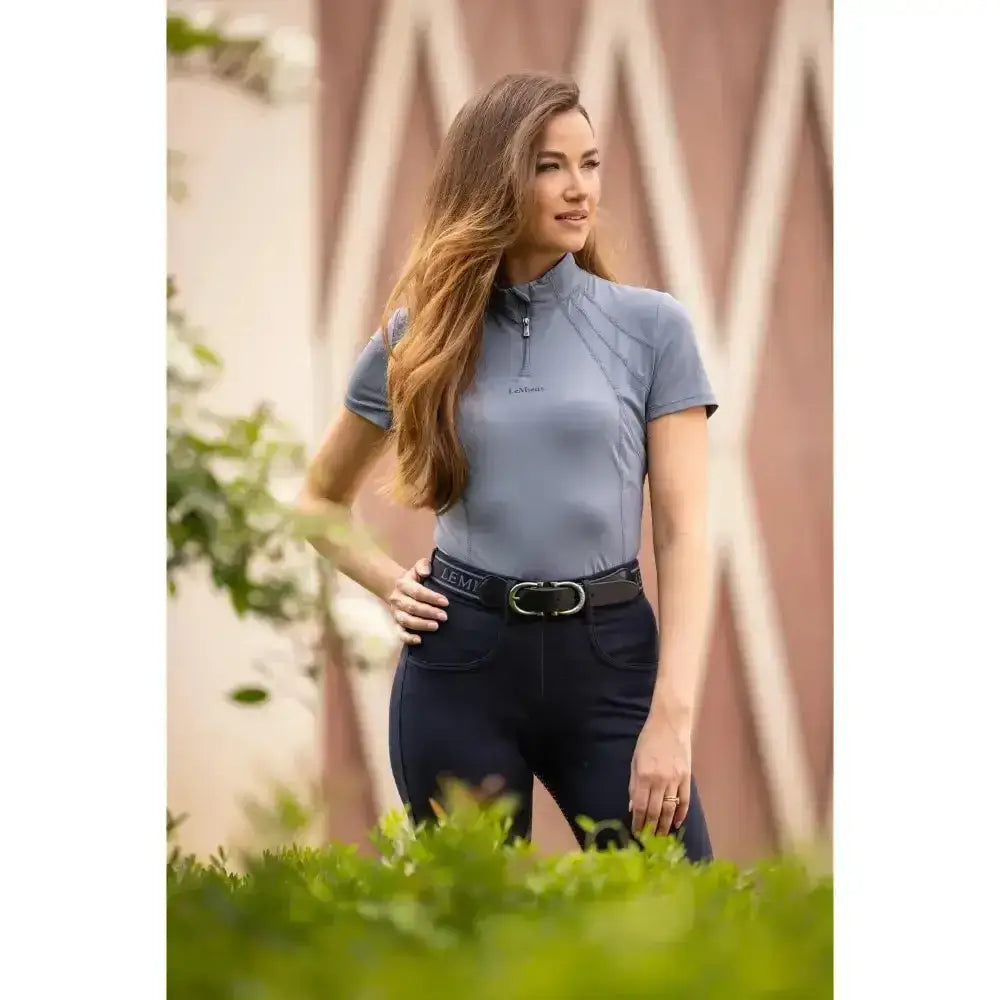 A woman wears a light blue lemieux mia mesh short sleeve base layer jay blue with a high collar and zipper detail