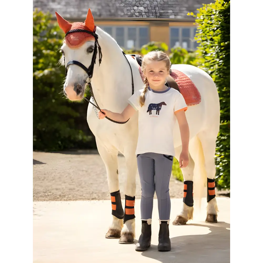 White horse with orange ears and striped leg wraps next to child in Lemieux Mini Alex Tee