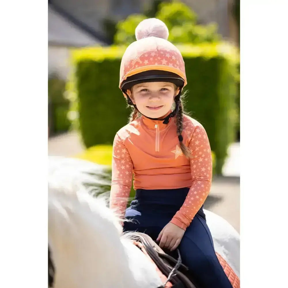 A child wearing a pink and orange equestrian helmet with pom pom and lemieux mini base layer apricot with ombre star print