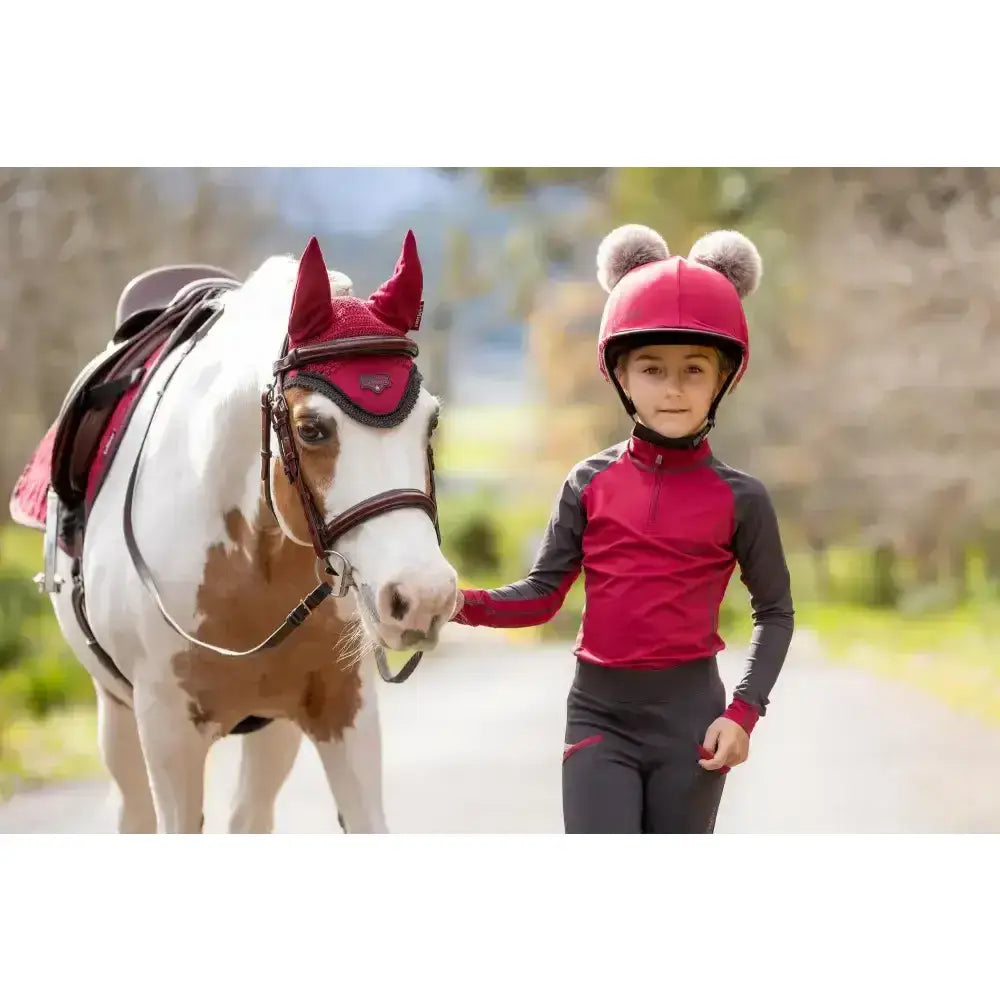 A young girl in a red and black lemieux mini base layer ember outfit stands by a white and brown horse in red gear