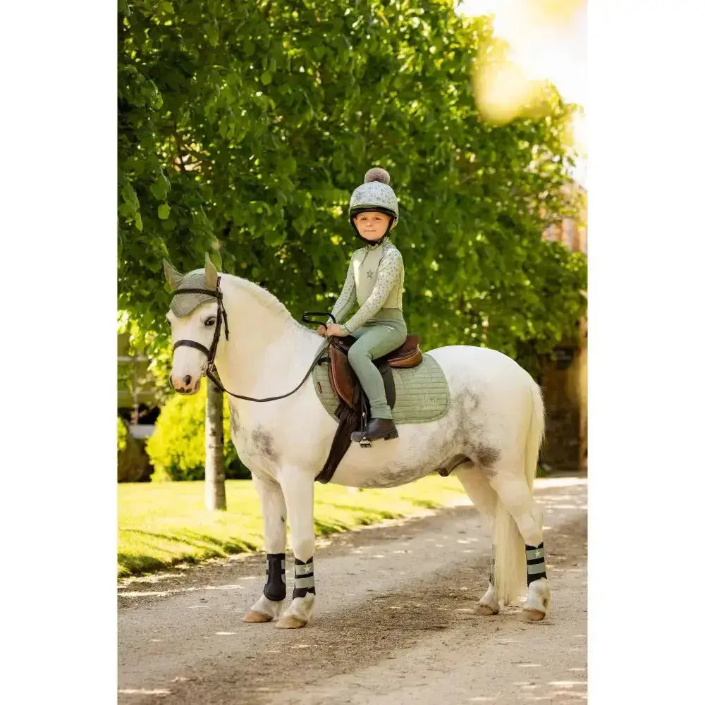 A young rider in a light green lemieux mini base layer fern outfit sits on a white horse ready for mini riders running
