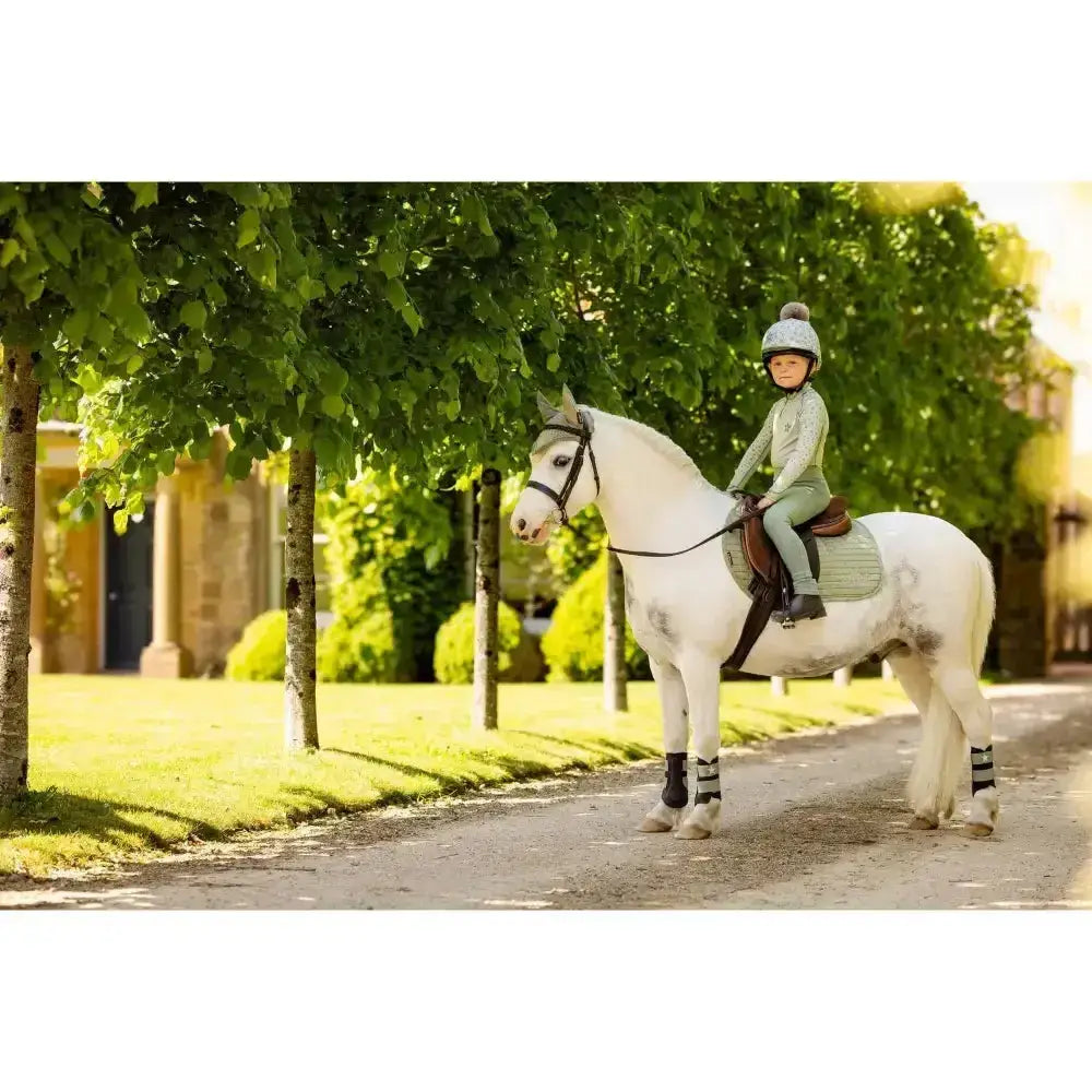 A young rider wearing the lemieux mini base layer fern in light green sits on a white horse on a sunny path