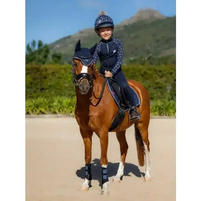 A young rider wears a navy blue lemieux mini base layer indigo top while riding a chestnut horse with protective gear