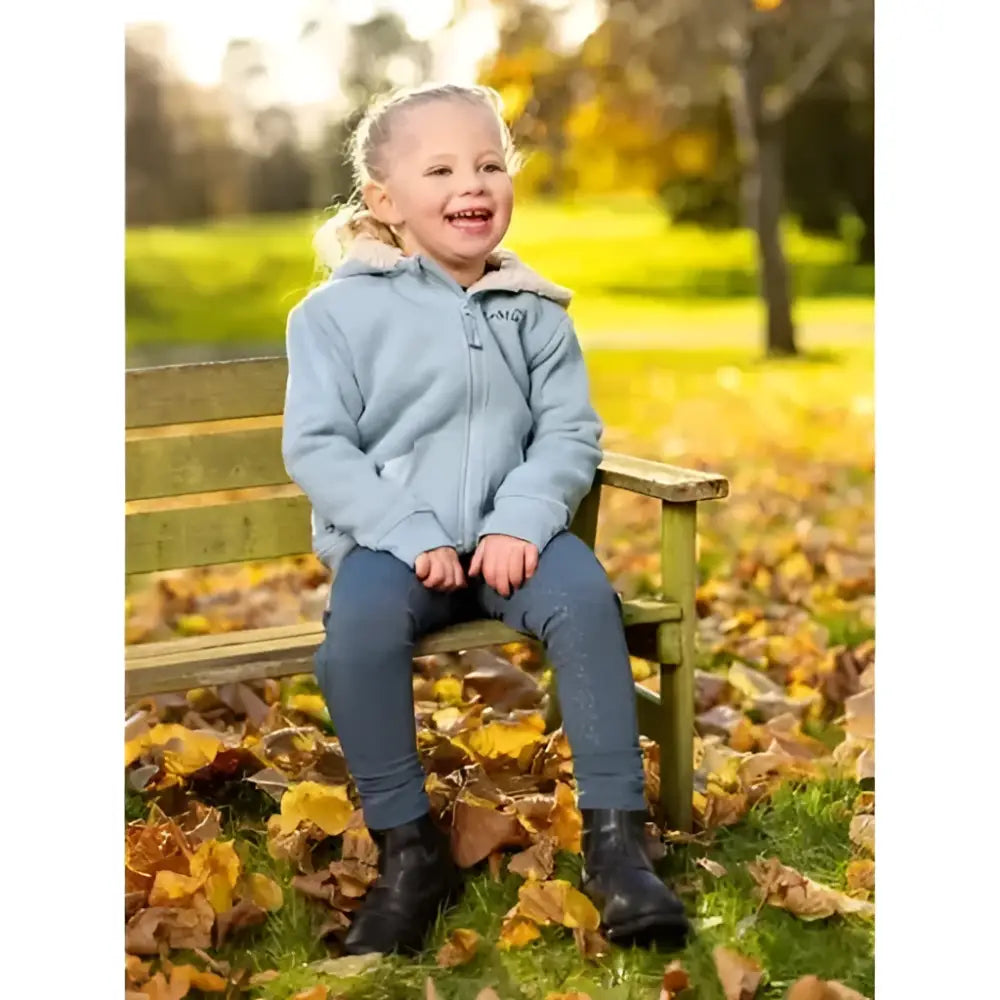 A young girl wearing a lemieux mini charlie super snuggly lined hoodie glacier sits on a wooden bench