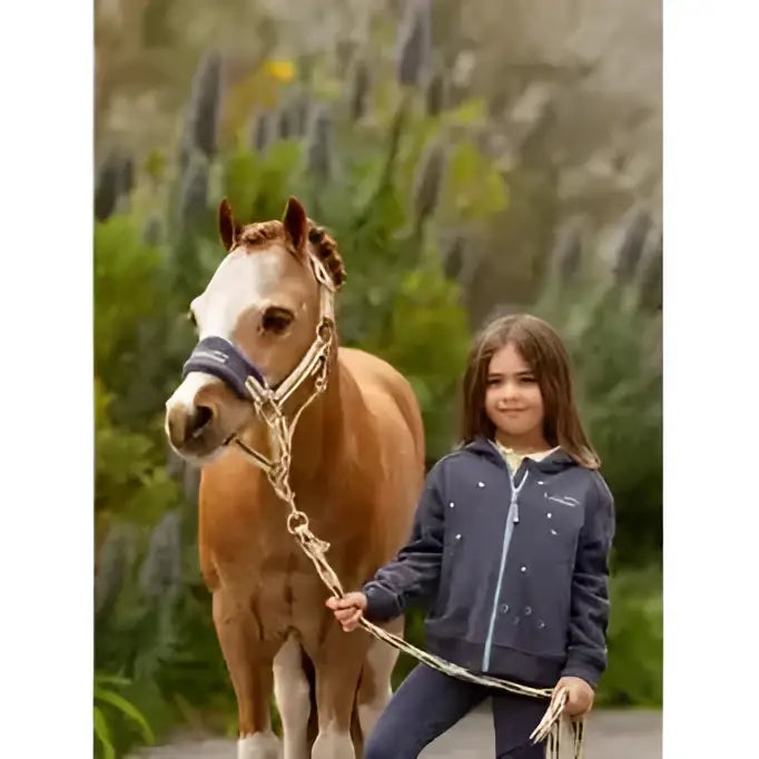 A young girl wearing a navy blue lemieux mini charlie zip jacket with silver stars stands beside a light brown horse