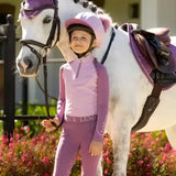 A young girl in a lavender lemieux mini double equestrian outfit stands beside a white horse with purple silk fondant gear and pom hat silk