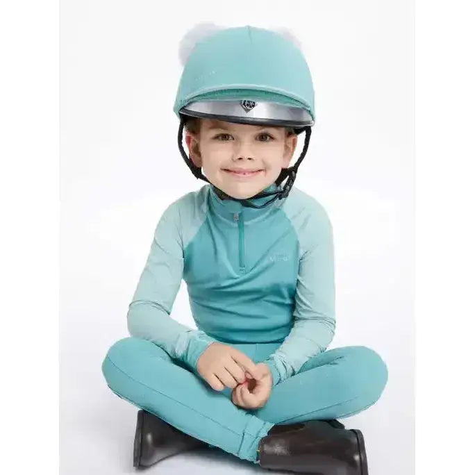 A child wearing a lemieux mini double pom double pom hat in silk lagoon teal with silver visor and white pom-poms