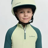 A child wearing a pale green and dark teal lemieux mini equestrian helmet with black chin strap and silk macaron faux fur pom