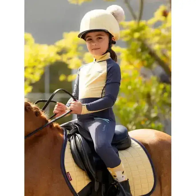 Child wearing a cream-colored lemieux mini double pom hat silk mimosa equestrian helmet with a fluffy pom-pom on top