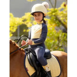 Child wearing a cream-colored lemieux mini double pom hat silk mimosa equestrian helmet with a fluffy pom-pom on top