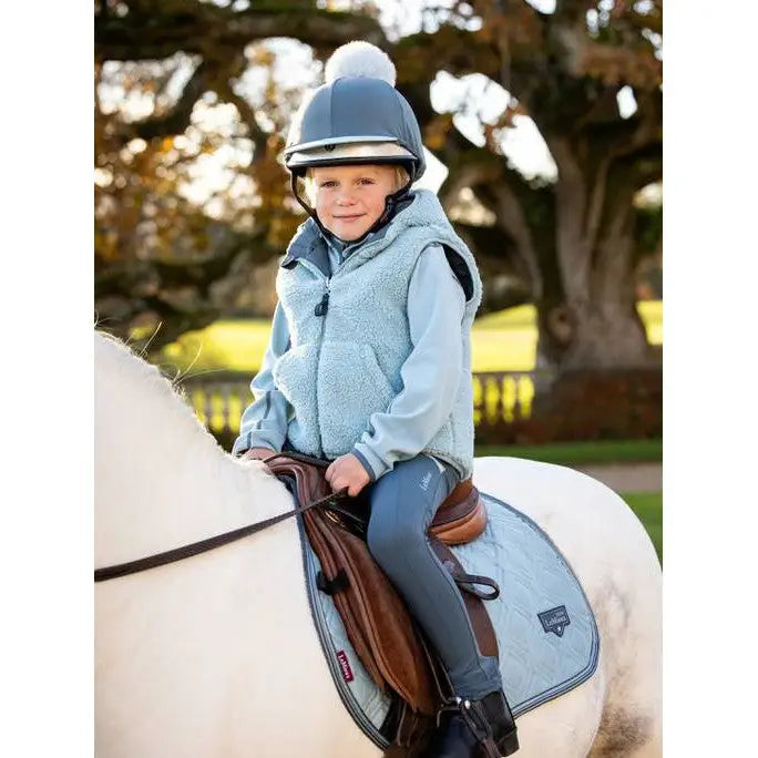 Young child in blue outfit and helmet riding white horse, featuring Lemieux Mini Elsie Gilet