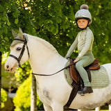 A young rider wearing a lemieux mini fly hood fern in green sits on a white horse with dark markings for riding