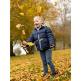 A young child joyfully stands in a navy reversible puffer navy jacket from lemieux mini megan surrounded by autumn leaves