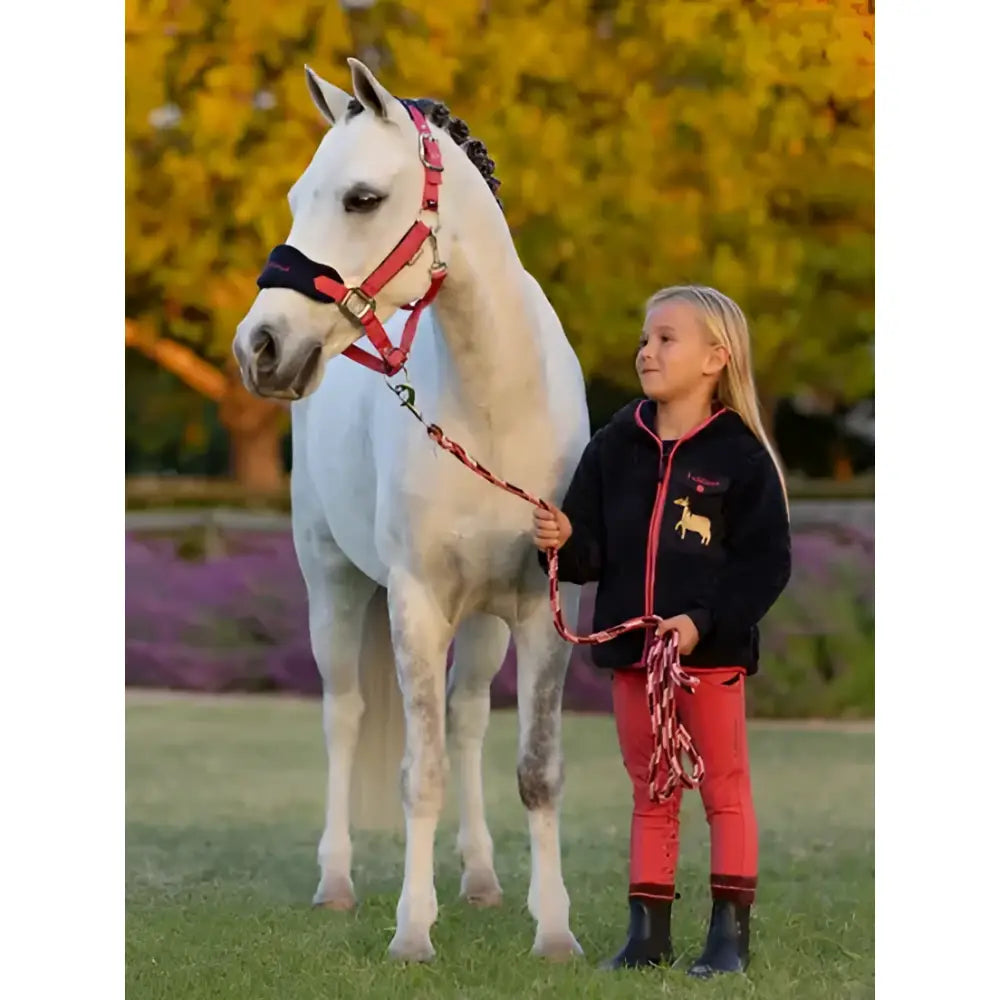 A white horse with a red halter and matching lead rope stands with a girl in an autumn field wearing lemieux mini pull ons cranberry