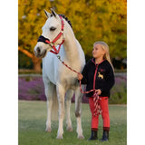 A white horse with a red halter and matching lead rope stands with a girl in an autumn field wearing lemieux mini pull ons cranberry