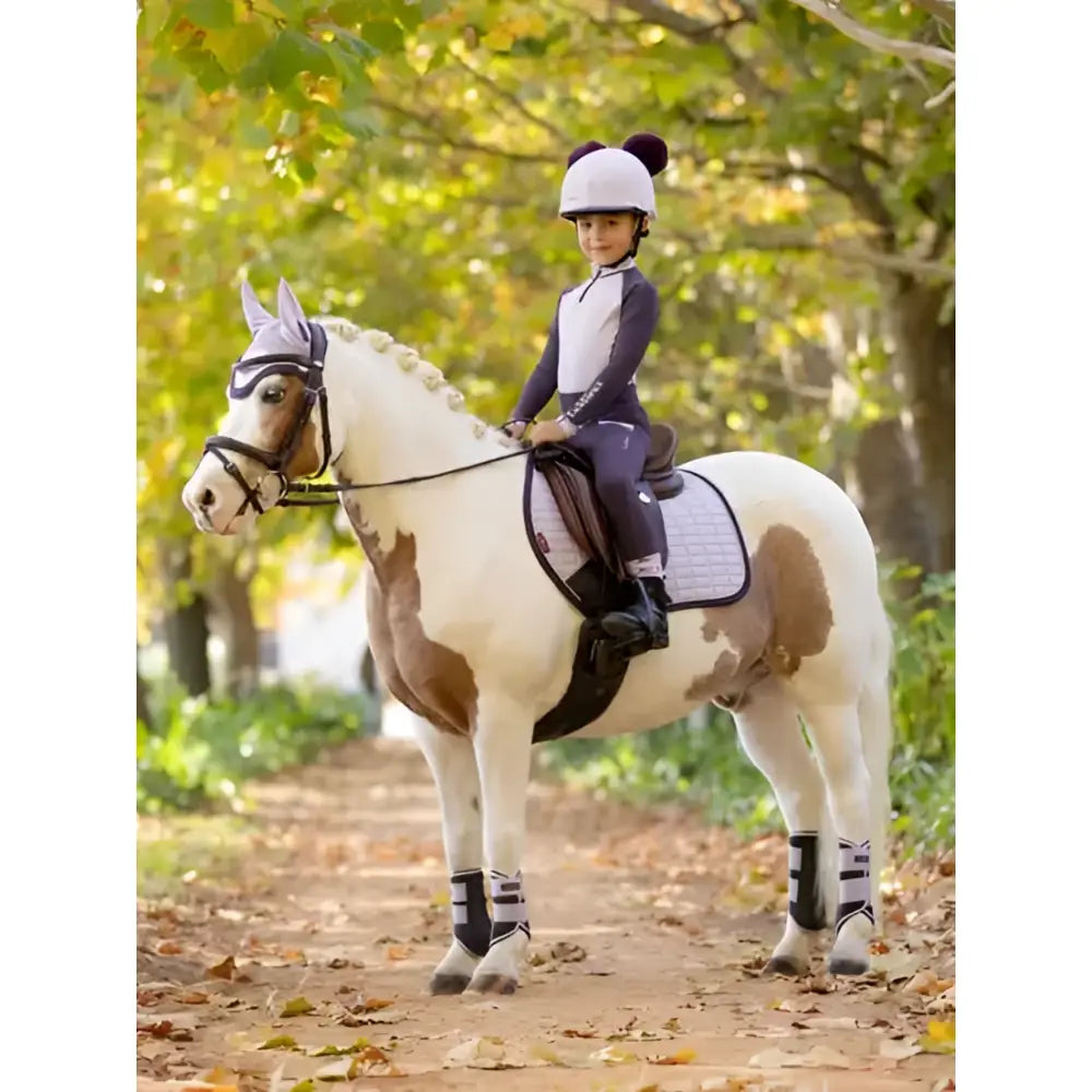 A young rider in a white helmet rides a pinto horse wearing lemieux mini pull ons juniper protective leg wraps