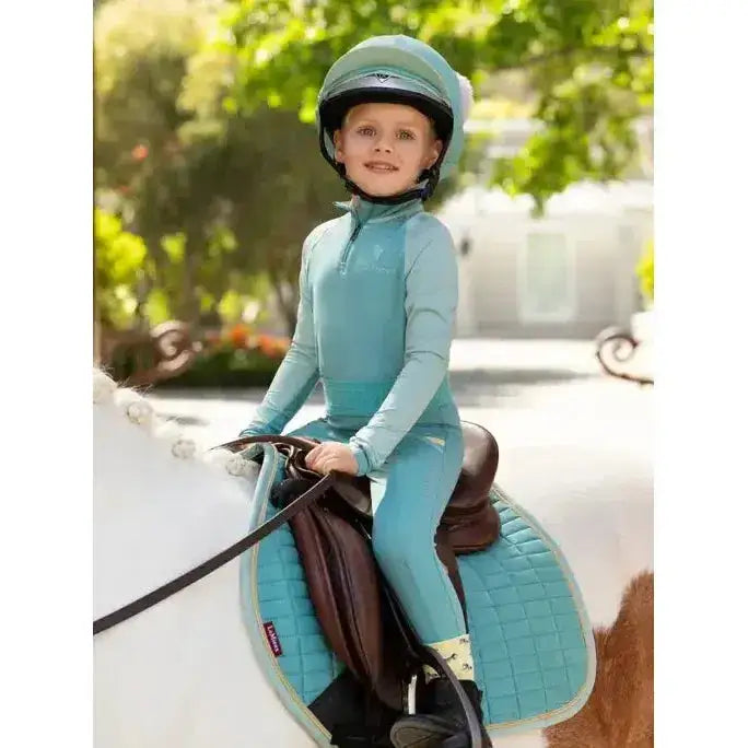 A young child in teal lemieux mini pull ons lagoon riding outfit and helmet seated on a white horse