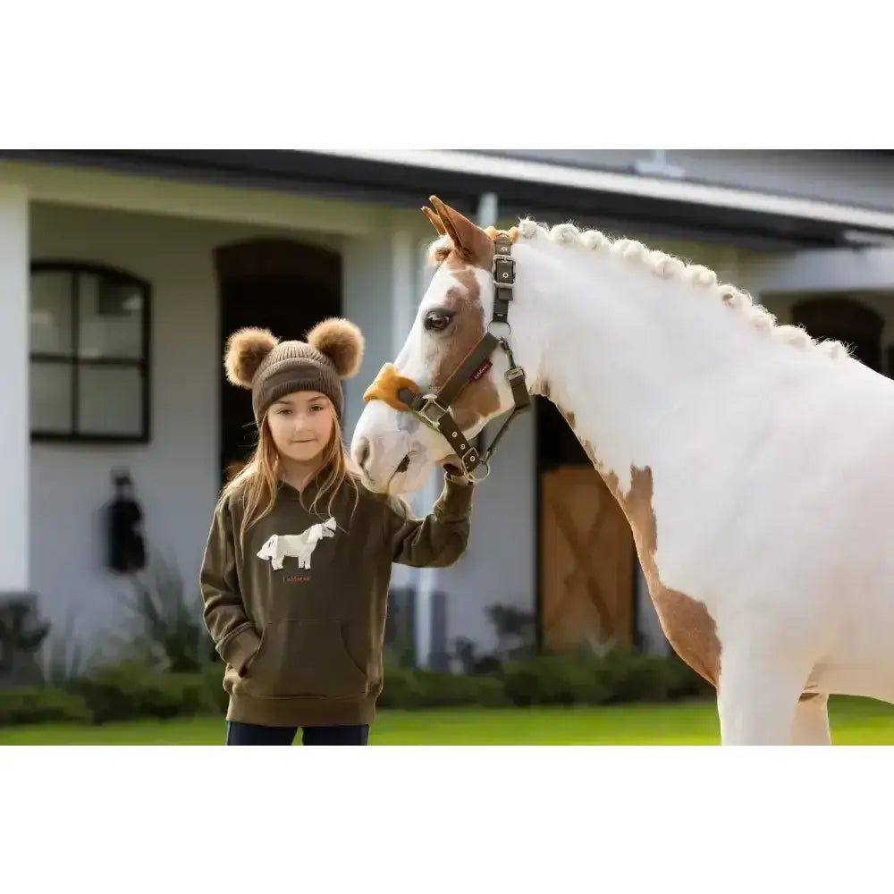 A young girl wearing a brown lemieux mini romi hoodie alpine with a white horse graphic and matching beanie with pom poms
