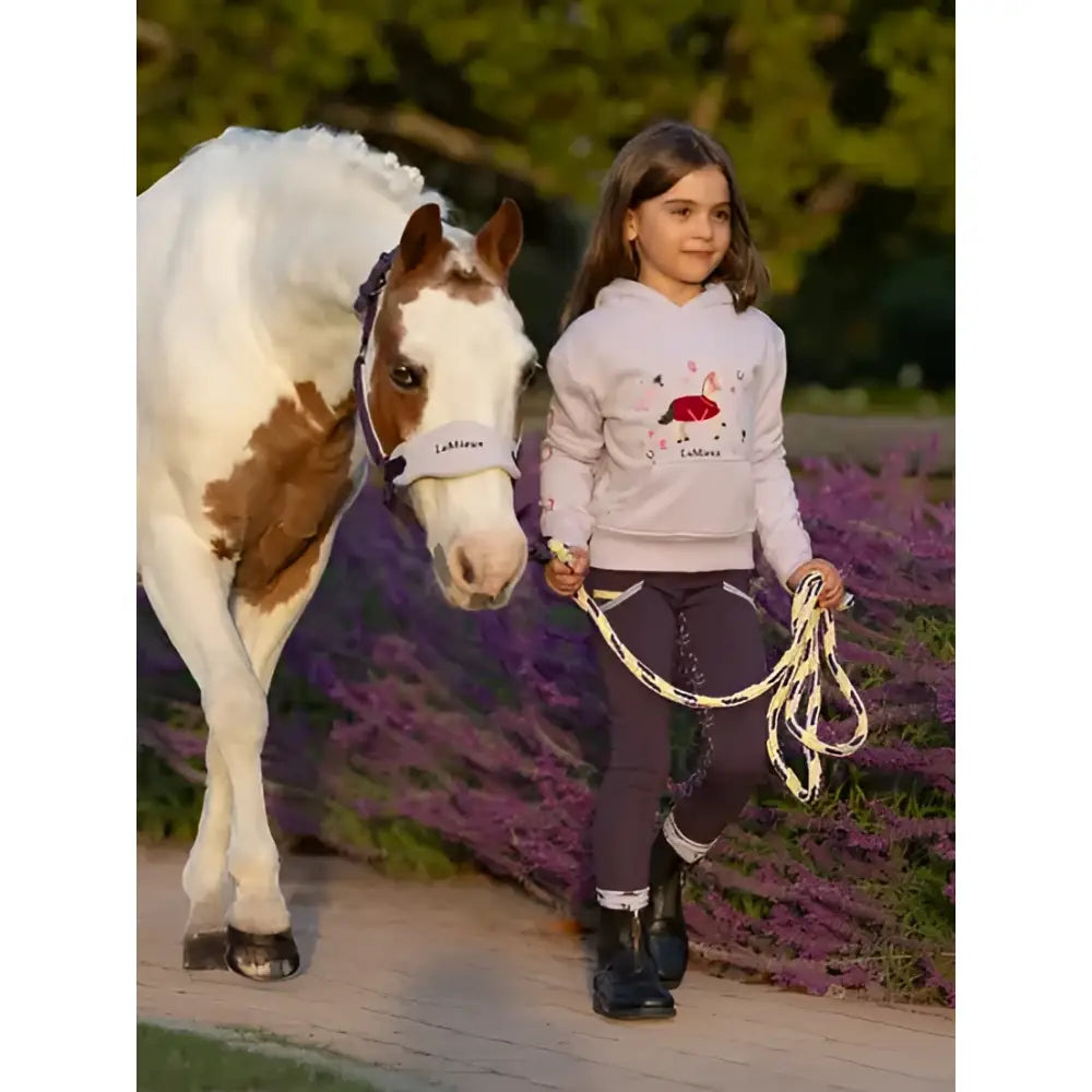 A young girl in a lemieux mini romi lilac hoodie with floral embroidery walks beside a white and brown horse