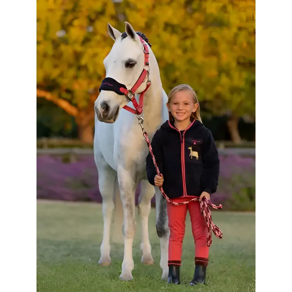 A young girl wearing lemieux mini teagan dream fleece navy stands beside a white horse holding its red lead rope