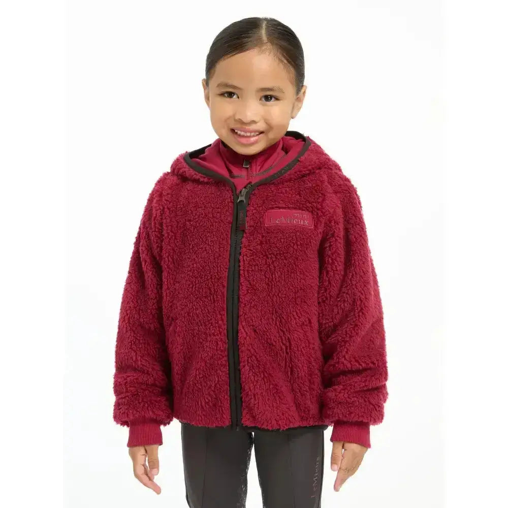 A child wearing a maroon lemieux mini teagan fleece ember jacket with black zipper and hood trim made of plush fleece