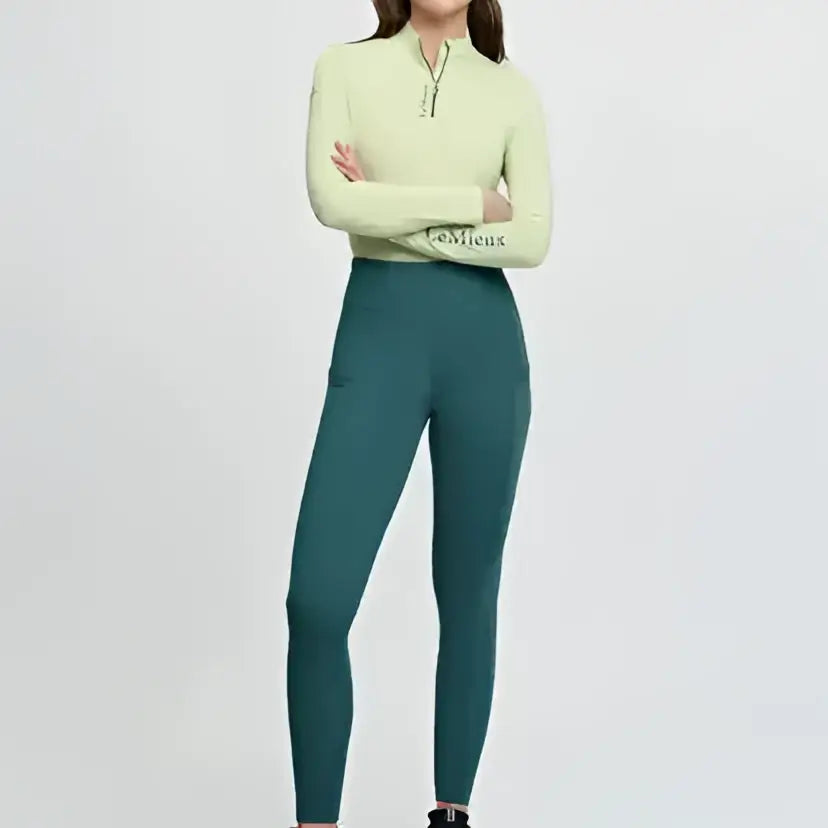 A woman wears a light green lemieux naomi pull zip neck top with dark teal matching leggings in the naomi pull breech jungle