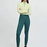 A woman wears a light green lemieux naomi pull zip neck top with dark teal matching leggings in the naomi pull breech jungle