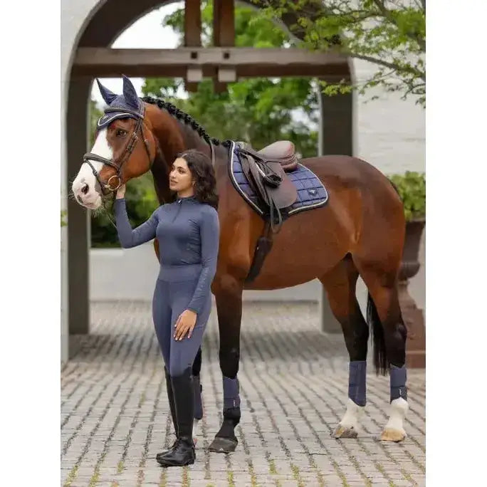 A brown horse with white markings wearing dark blue protective gear paired with lemieux naomi pull breeches dusk blue