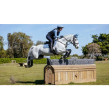 A white horse with black hooves leaps over a wooden obstacle wearing lemieux suede close contact square ice blue bridle