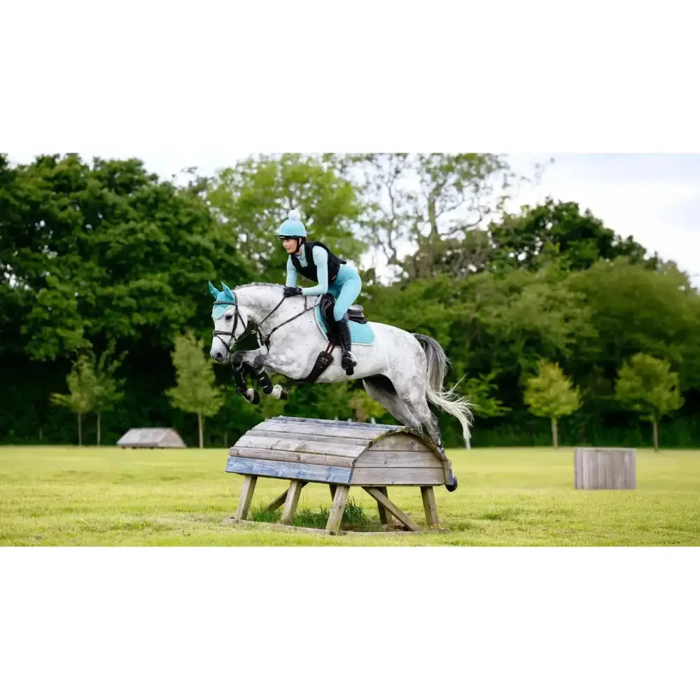 A white horse wearing lemieux suede close contact square lagoon teal protective gear mid-jump with matching rider