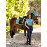 A woman stands beside a brown horse wearing teal lemieux suede dressage square lagoon leg wraps and saddle pad