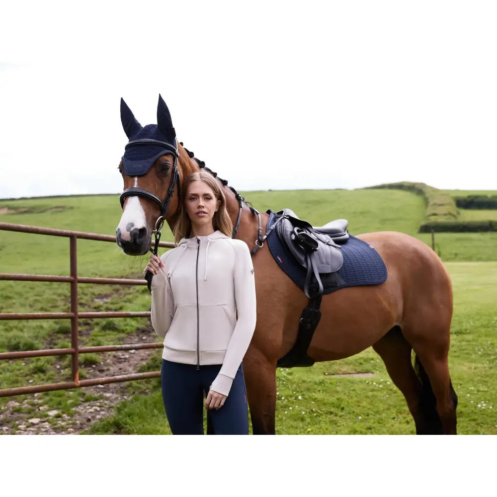 A woman wearing a lemieux tabi tech zip through hoodie stone stands beside a brown horse with black riding gear