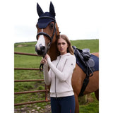 A woman in a lemieux tabi tech stone hoodie zip stands beside a horse wearing a navy fly mask in outdoor setting