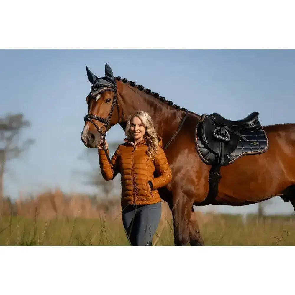 Brown horse stands in a field wearing a black bridle and saddle next to a lemieux tilly hooded puffer jacket ginger display