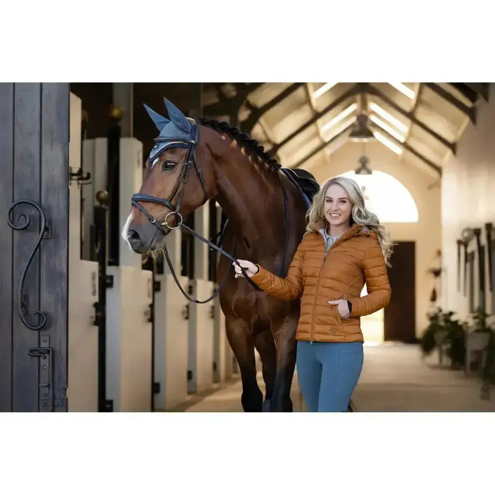 A horse wearing a blue pointed ear cover alongside a lemieux tilly hooded puffer jacket ginger in puffer jacket ginger color