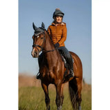 A woman wearing a lemieux tilly hooded puffer jacket ginger rides a glossy brown horse with black tack and headgear