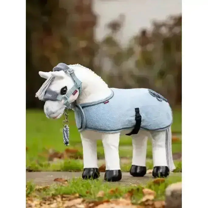 A plush white lemieux toy pony coco wearing a light blue textured blanket with black straps and a gray bridle